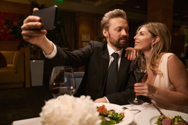 Zarif bir çift restoranda selfie çeker, gece elbisesi giymiş bir kadın.