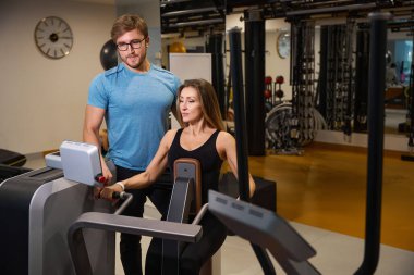 Kadın, bir eğitmenin gözetiminde kaslarını ısıtmak için bir makinede çalışıyor.