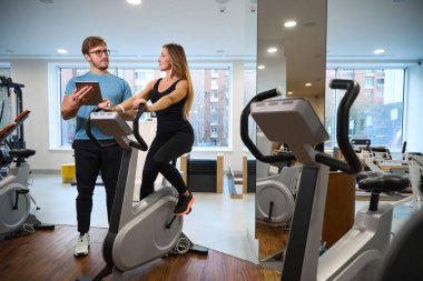 Eğitmen bir kadınla egzersiz bisikleti üzerinde iletişim kuruyor, odada bir sürü egzersiz makinesi var.