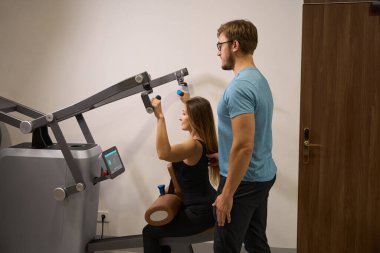 Sağlık merkezindeki kadın müşteri, bir eğitmenin denetimi altında bir güç makinesinde çalışır.