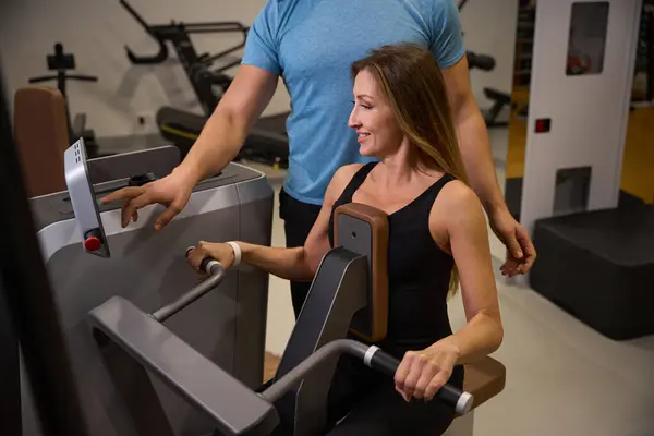 Middle-aged lady is working out on machine under the supervision of a trainer, there is modern equipment in the gym