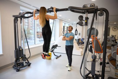 Spor salonunda bir eğitmenin gözetiminde barfiks çeken bir kadın.