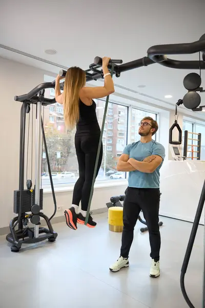 Lady in the gym doing pull-ups on the horizontal bar under the supervision of an instructor
