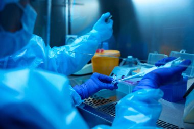 Laboratuvar çalışanları işlerinde pipet makinesi kullanıyorlar. Masada üzerinde biyomateryal olan sarı bir kap var.