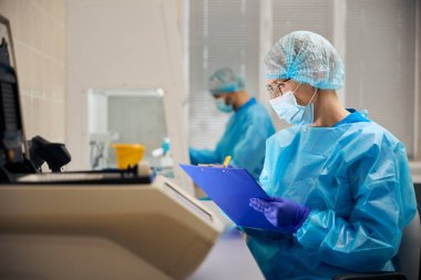 Kadın laboratuvar asistanı araştırma sonuçlarını kaydediyor, meslektaşı da yakınlarda çalışıyor.