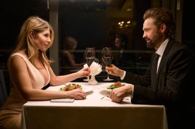 Akşam elbisesi giymiş bir kadın ve şık bir iş elbisesi giymiş bir adam, rahat bir restoranda akşam yemeğinde konuşuyor.