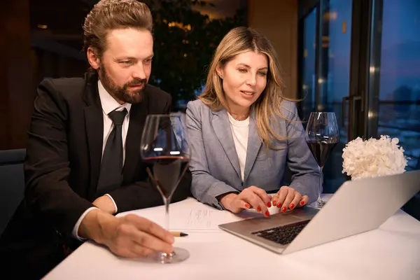 Man and a woman in business suits sit in a restaurant with a laptop, glasses of wine on the table