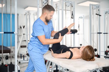 Mavi üniformalı bir adam rehabilitasyon merkezinde bir hastayla çalışıyor. Rahat bir eşofman giyen bir kadın.