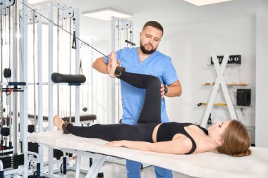 Deneyimli fizyoterapist bir hastayla çalışmak için özel bir egzersiz makinesi kullanır, kadın masaj masasında yatar.