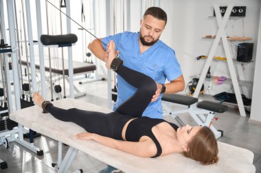 Deneyimli bir kinezolog hastayla çalışmak için özel bir simülatör kullanır. Kadın masaj masasında yatar.