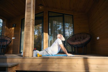 Gray-bearded old man is doing yoga in the fresh air, he is located on a wooden terrace clipart