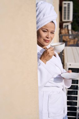 Elinde kahve fincanı ve balkon duvarına yaslanmış çay tabağıyla bornozlu olgun bir kadın.