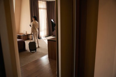 Tramvay valizi ve seyahat belgeleriyle otel odasının ortasında duran bayanın arka görüntüsü.