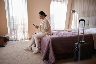 Otel odasında yatakta oturmuş akıllı telefon ekranına bakan odaklanmış kadın turist.