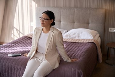 Gözlüklü, memnun iş kadını otel odasında yatağın kenarına oturmuş başka tarafa bakıyor.