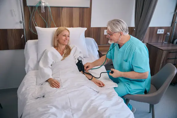 女性住院病人躺在病床上微笑 同时在病房里看医生 测量血压 — 图库照片