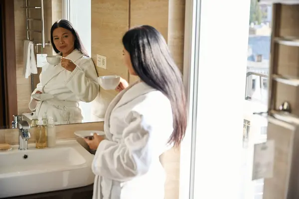 バスルームの壁に傾いている間 鏡で自分自身を見つめる手でカップとソーサーでバスローブに満足した女性 ロイヤリティフリーのストック写真
