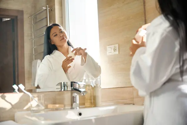 Bornozlu Kadın Aynadaki Yansımasına Bakarken Kozmetik Krem Kavanozunun Kapağını Açıyor Stok Resim
