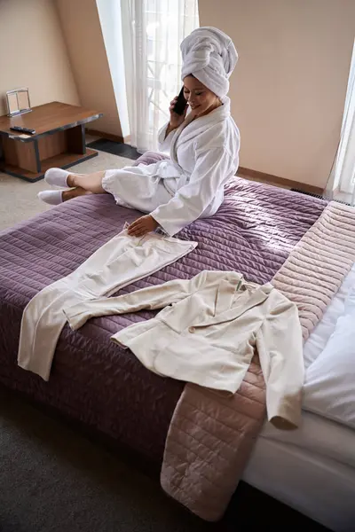 在电话交谈中 穿着浴衣的心满意足的女士坐在酒店房间的床上 看着她的西服裤 图库图片