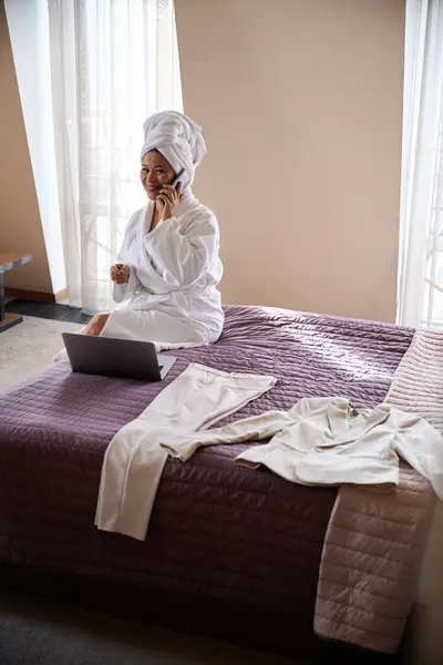 在电话交谈中 穿着浴衣的快乐的女商人坐在她的手提电脑前的豪华套间里 图库图片