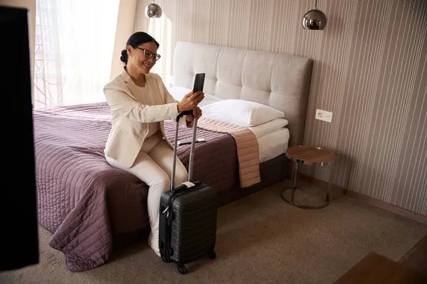 Lächelnde Frau Sitzt Hotelzimmer Auf Dem Bett Und Lehnt Reisekoffergriff Stockbild