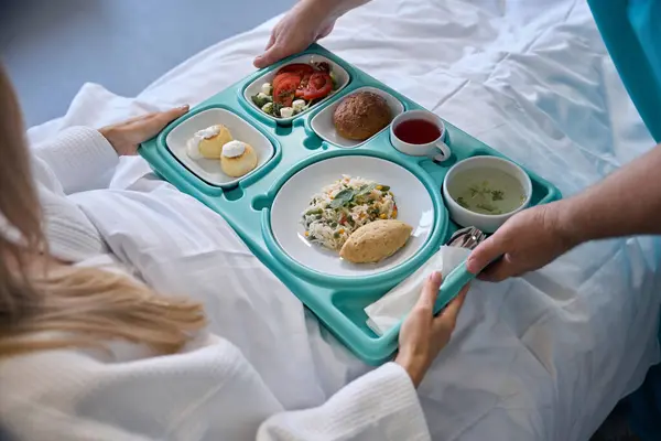 病院のベッドに座っている女性の入院患者に食品トレイを与える男性看護師の手の汚れた写真 ストック写真