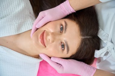 Kozmetikçi ellerini nitrile eldivenlerle yanaklarına masaj yaparken sırtüstü uzanan genç bir kadının en iyi görüntüsü.