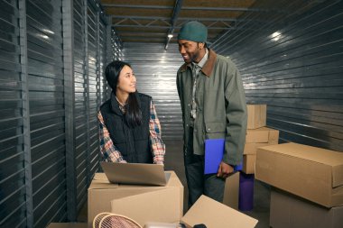 Gülümseyen yönetici, karton kutuların arasında çelik konteynırın içinde dizüstü bilgisayardaki bir şeyi depo çalışanına gösteriyor.