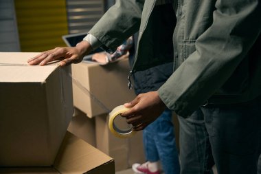 Cropped photo of storehouse employee sealing cardboard box with packing tape in storage container while his coworker typing on laptop clipart