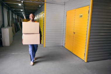 Modern depoda bir çift karton kutu taşıyan memnun yükleyicinin tam boy portresi.