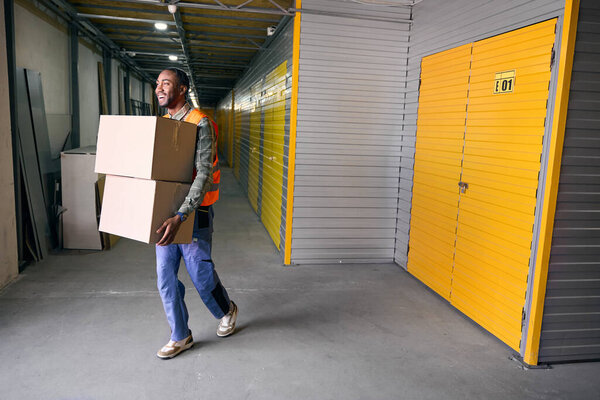 Full-size portrait of merry freight handler carrying pair of cardboard boxes in storage area
