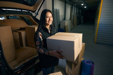 Güzel bir kadın bir deponun bagajında otururken elinde bir kutu tutuyor.