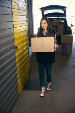 Gülümseyen Asyalı kadın bir arabanın arka planında elinde bir kutu tutuyor. Tam uzunluk