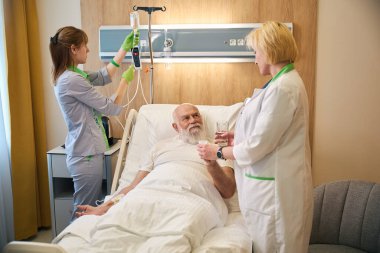 Yaşlı hasta hastane yatağında yatarken doktorla iletişim kuruyor.