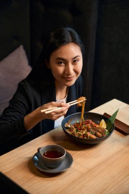 Yemek çubuklarıyla erişte tutan, kameraya bakan, lezzetli ve lezzetli deniz mahsullü ramen yiyen, iştahlı ve zevkli Asyalı kadın, hayatın tadını çıkarıyor.