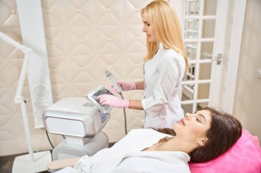 Kadın kanepede yatarken, deri uzmanı azot eldivenleri ile RF mikron ayarlarını ayarlıyor.