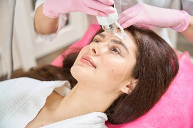 Güzellik prosedürü sırasında estetik tıp kliniği müşterisi koltuğa uzanmış kozmetik uzmanı mikronedle RF el yazısını alnına yerleştiriyor.