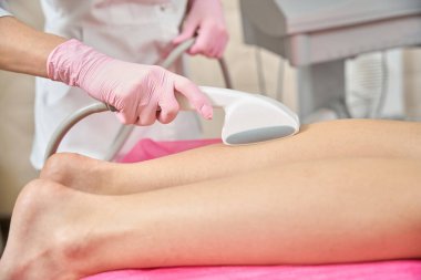 Tek kullanımlık nitrile eldivenli estetikçinin kesilmiş fotoğrafı hasta baldırının üzerinde güzellik aletini hareket ettiriyor.