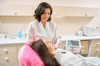 Konsantre güzellik uzmanı koltukta sırtüstü uzanan kadın hastanın boynuna mikronedle radyo frekansı el yazısı bastırıyor.