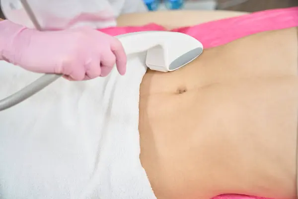 stock image Closeup of hand in nitrile glove pressing beauty machine handpiece against abdomen of female client in supine position