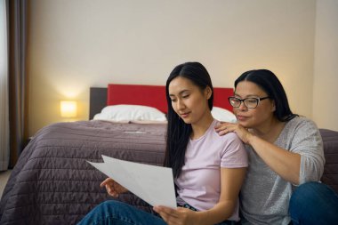 Odaklanmış genç bir kadın ve annesi belgelerini okurken yatağın kenarında oturuyorlar.