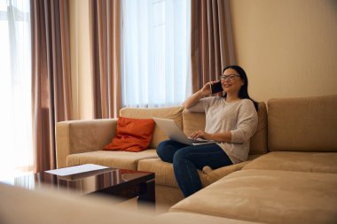 Portatif bilgisayarlı gülümseyen bayan köşedeki koltukta oturmuş cep telefonuyla konuşuyor.