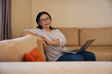 Elinde taşınabilir bilgisayar ve cep telefonuyla rahat koltukta oturan Asyalı kadın geleceğe bakıyor.