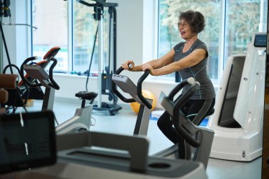 Gülümseyen yaşlı kadın egzersiz bisikletinde egzersiz yapıyor, bir fitness kliniğinde rehabilitasyona gidiyor.