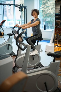 Gülümseyen yaşlı bayan egzersiz bisikletinde egzersiz yapıyor, bir fitness kliniğinde rehabilitasyona gidiyor.