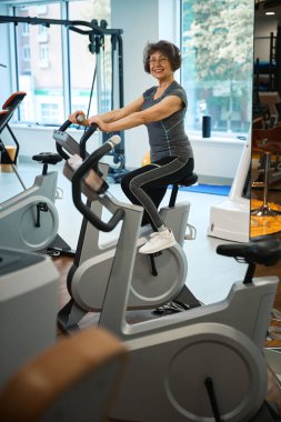 Smiling elderly lady is exercising on an exercise bike, she is undergoing rehabilitation in a fitness clinic clipart