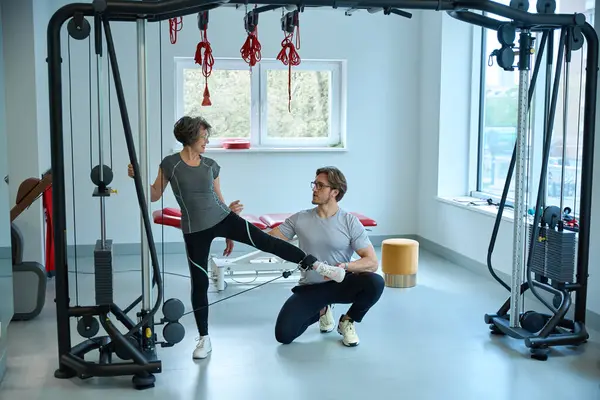 stock image Young trainer is working with an elderly patient on a special simulator, the gym has modern equipment