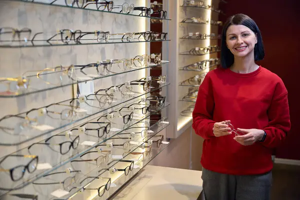 stock image Brunette stands in an optician near a display case with glasses, she is wearing a red sweatshirt