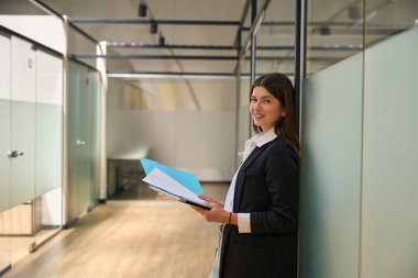 Bir ofis binasının koridorunda duran ve kameraya bakan genç bir kadın.