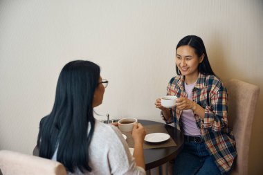 Neşeli genç bayan ve annesi ellerinde kahveyle yuvarlak masada oturuyorlar.